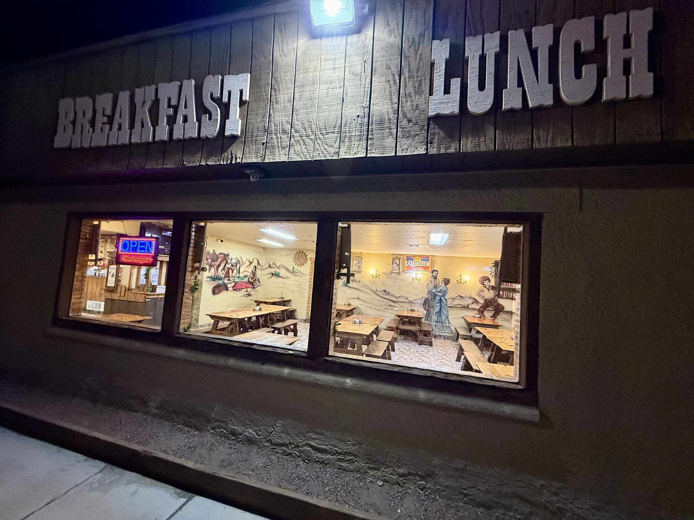 The Wild West Cowboy Steakhouse storefront at night, the painted Western window murals lit from inside with picnic tables visible and an Open sign.